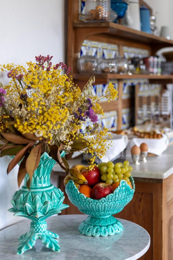A turquoise vase with yellow and purple flowers and a matching fruit bowl filled with apples, grapes, and bananas sit on a marble table—evoking the inviting ambiance of hotels in Cantabria—with a kitchen and breakfast items in the background.