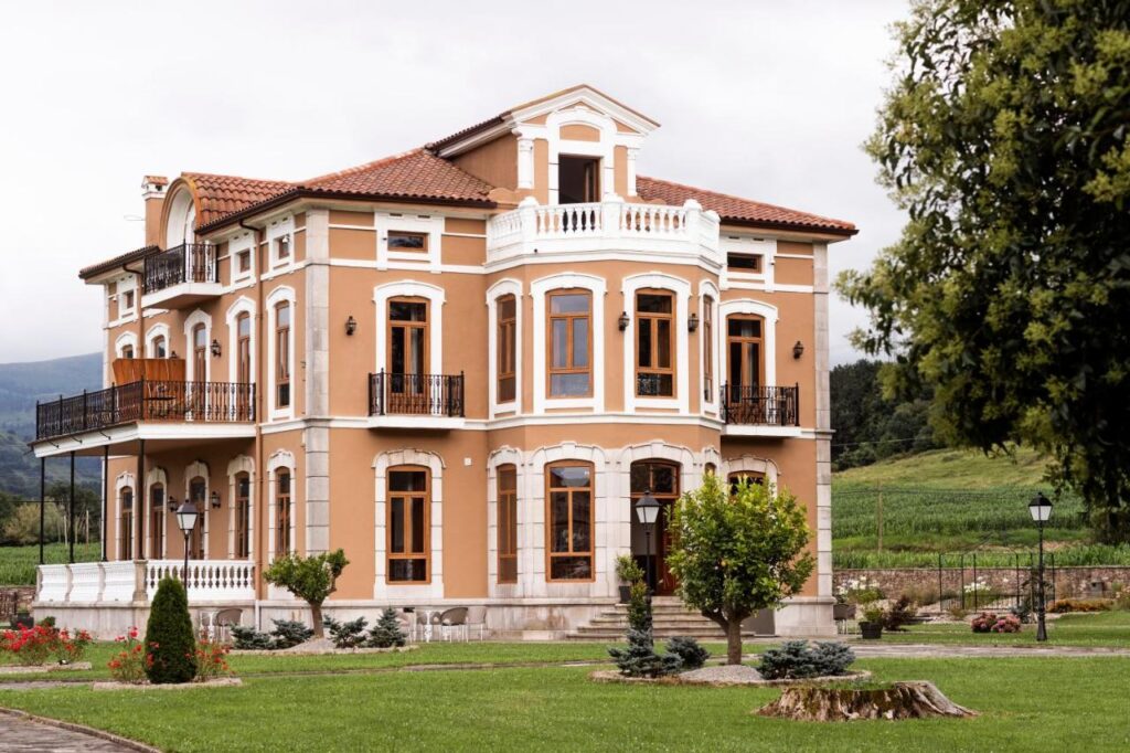 A large, elegant, three-story house with tan walls, white trim, and multiple windows—reminiscent of boutique hotels in Cantabria—surrounded by a well-maintained lawn, trees, shrubs, and a garden, with hills and greenery in the background.