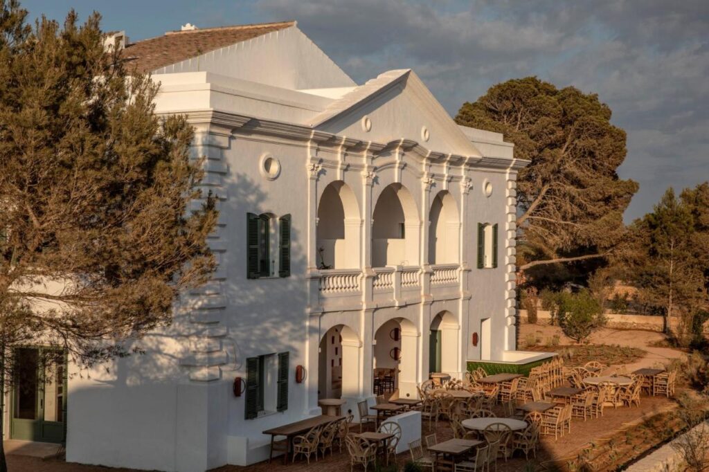 Unique Boutique Hotels in Menorca 22 A large white Mediterranean-style building with arched balconies, green shutters, and outdoor seating, surrounded by trees and bathed in warm sunlight—an inviting example of boutique hotels in Menorca.