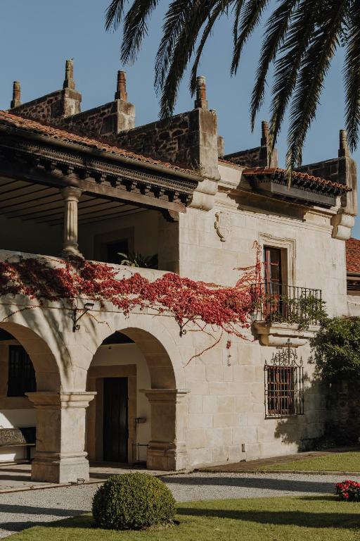 A historic stone building with arched doorways, wrought iron balcony, and red roof tiles, this charming spot is reminiscent of the elegant hotels in Cantabria. Red vines climb the walls as palm tree leaves sway above neat trimmed bushes.