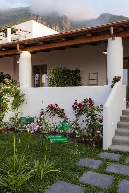 7 Best Hotels in the Aeolian Islands 10 A white stucco house with a wooden roof sits in front of lush green mountains, resembling the charming hotels in the Aeolian Islands, and features a small garden with flowers and stone paths leading to steps up to the porch.