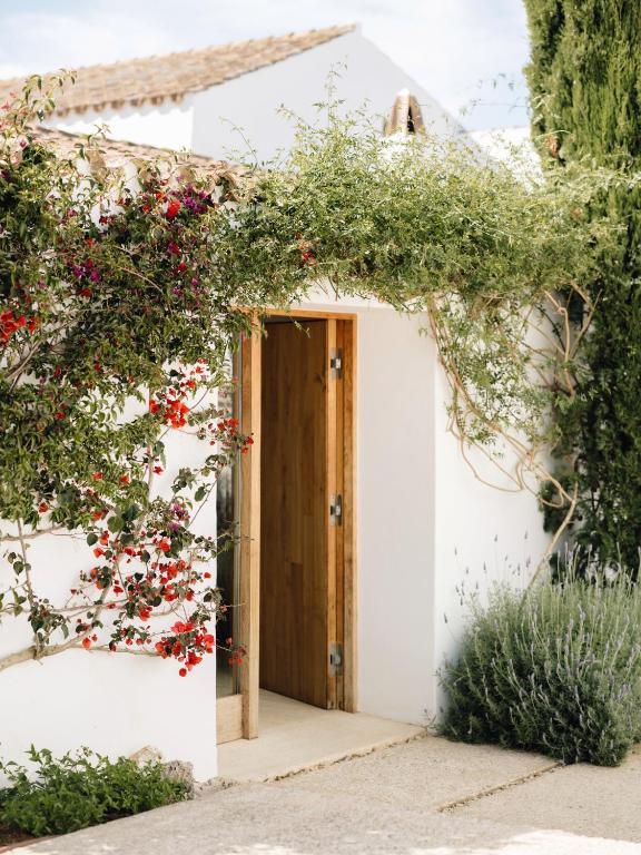 Unique Boutique Hotels in Menorca 38 A wooden door set into a white stucco wall is surrounded by lush greenery and blooming red flowers, with lavender plants and tile-roofed buildings reminiscent of boutique hotels in Menorca visible in the background.