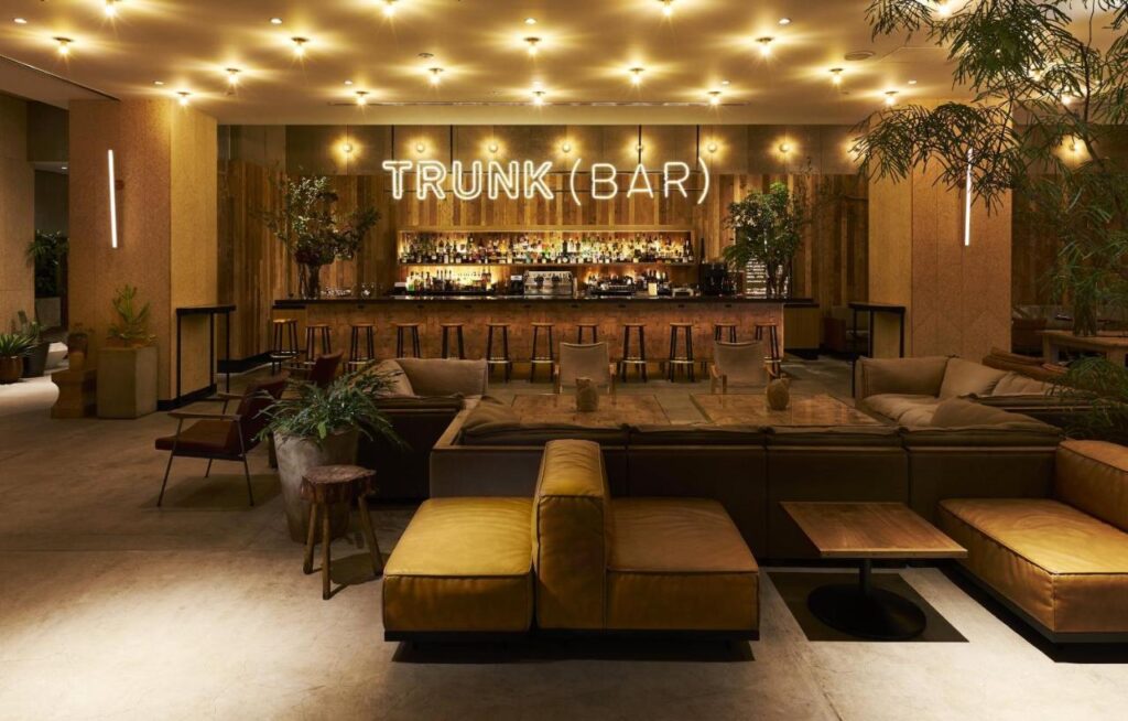 15 Unique Hotels in Tokyo: Coolest hotels 8 A stylish, modern bar called TRUNK(BAR), found in one of the unique hotels in Tokyo, features cozy brown sofas, wooden tables, indoor plants, and a well-lit bar with high stools and a wide selection of bottles behind the counter.