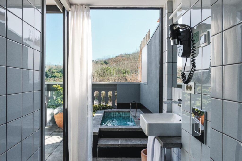 13 Unique Hotels in Barcelona, Spain 40 A modern bathroom with white tiles features a sink and mounted hairdryer, and opens onto a balcony with a small private pool and scenic views—perfect for those seeking unique hotels in Barcelona amid hills and trees through large glass doors.