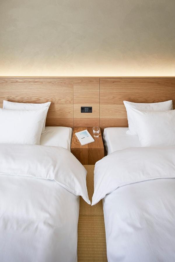 15 Unique Hotels in Tokyo: Coolest hotels 25 Two neatly made single beds with white bedding are separated by a wooden nightstand holding a glass of water and a closed book. This warm, softly lit room reflects the inviting charm found in many unique hotels in Tokyo.
