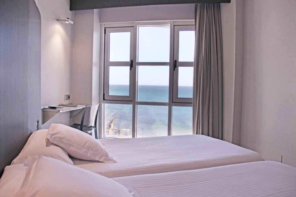 Two neatly made single beds in a bright room with large windows offering a clear view of the sea and sky. Perfect for guests seeking hotels in Cantabria, with a desk by the window and natural light filling the space.