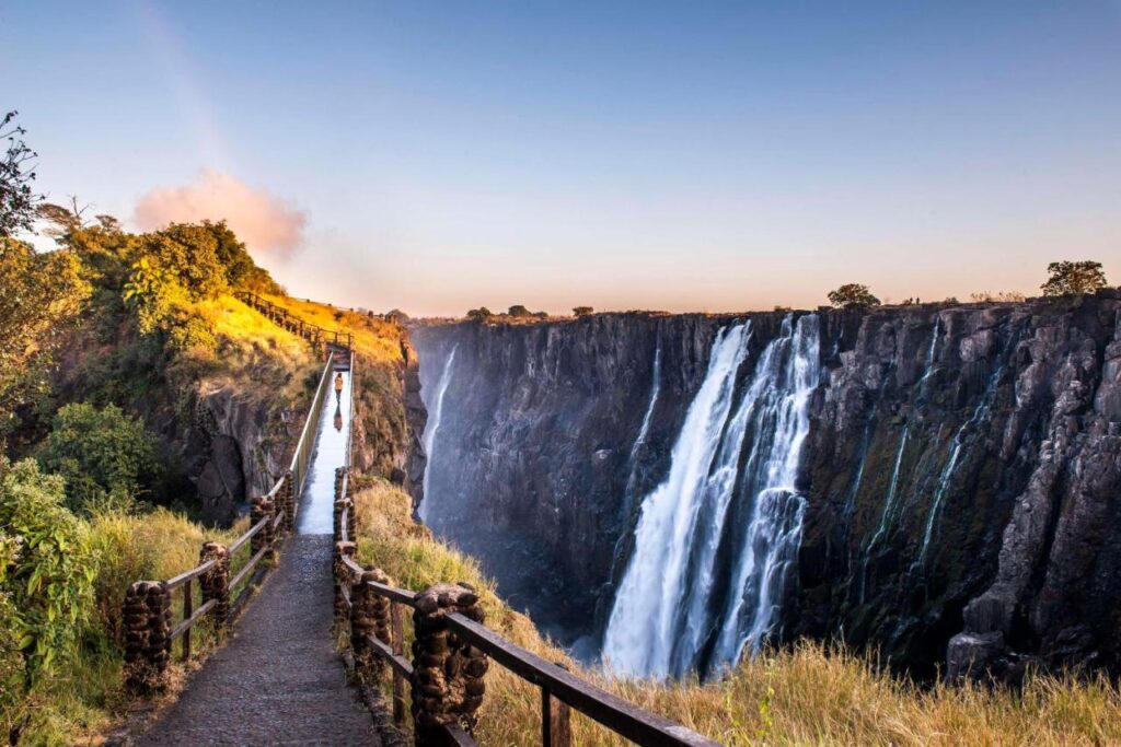 Best Hotels in Victoria Falls: Unique Luxury & Safari Stays 2 A scenic view of Victoria Falls with a narrow footbridge leading toward the waterfall, surrounded by lush greenery and dramatic cliffs under a clear blue sky—just steps away from some of the best hotels in Victoria Falls.