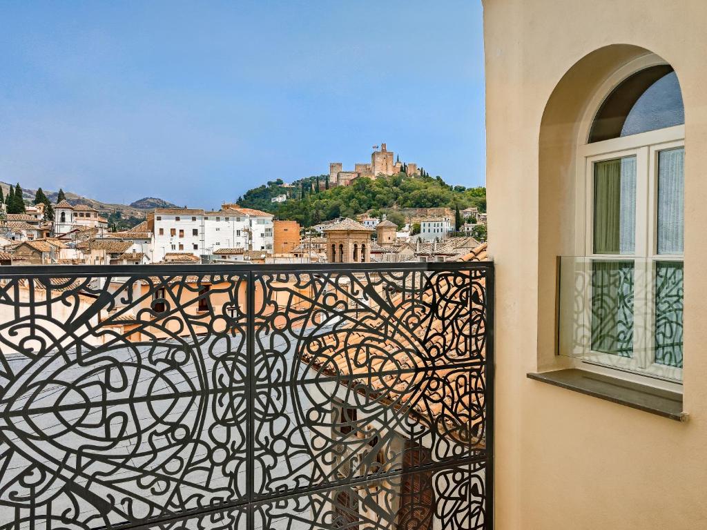 A cityscape view with decorative black wrought iron balcony railing, light-colored buildings, and a hilltop fortress in the distance under a clear blue sky—reminiscent of charming boutique hotels in Granada. An arched window is visible on the right.