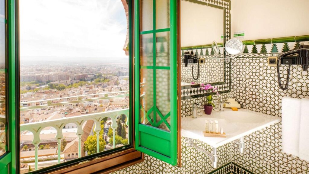 A bathroom with green and white patterned tiles, a corner sink, toiletries, and a purple orchid in a pot. An open window offers a panoramic city view, evoking the charm of boutique hotels in Granada beneath a bright sky.