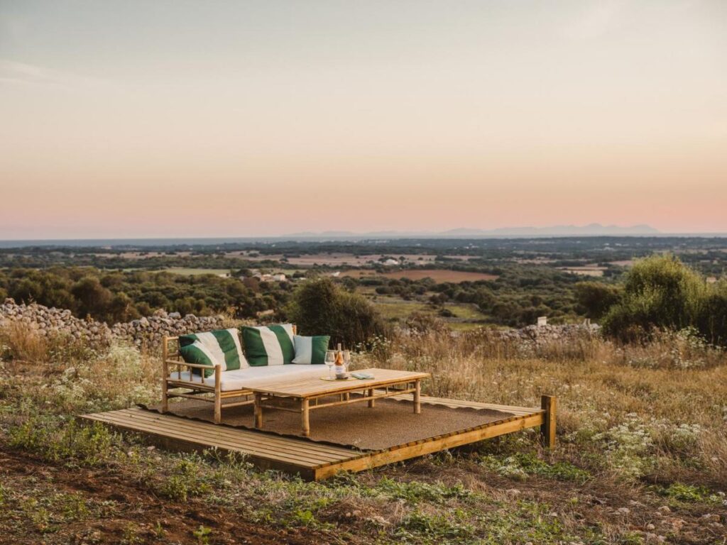 Unique Boutique Hotels in Menorca 12 A wooden outdoor sofa with green and white cushions and a table sits on a small deck in a grassy field, evoking the relaxed charm of boutique hotels in Menorca, overlooking vast landscapes at sunset with distant hills under a calm sky.