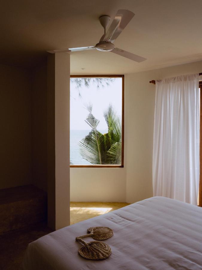 Unique Places to Stay in Sri Lanka: Boutique & Beach Hotels 2 A serene bedroom with white walls and bedding, a ceiling fan, a large window framing palm leaves and ocean views—sunlight streams through sheer curtains. Two woven fans rest on the bed in one of the unique places to stay in Sri Lanka.