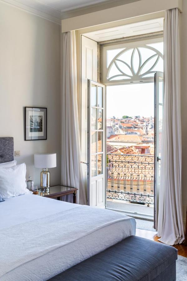 Top 12 Boutique Hotels in Porto, Portugal 36 A bright bedroom in the style of boutique hotels in Porto features a neatly made bed, bedside table, lamp, and framed picture. French doors open onto a balcony with ornate railing and views of city rooftops under a clear sky.