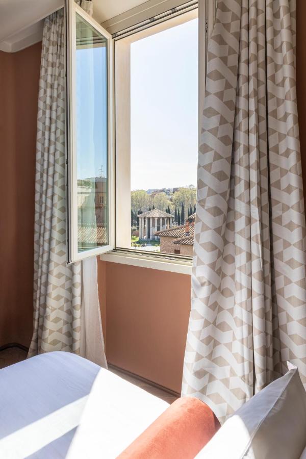 13 UNIQUE Boutique Hotels in Rome 19 A sunlit room in one of the boutique hotels in Rome, Italy, with white and patterned curtains drawn open, reveals a window view of an ancient Roman-style temple surrounded by trees and historic buildings.