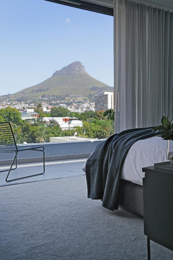 Unique Places to Stay in Cape Town 14 A modern bedroom with a large window offers a scenic view of a distinctive, cone-shaped mountain and cityscape—one of the unique places to stay in Cape Town. The room features a bed with a dark blanket, a chair, and sheer curtains.