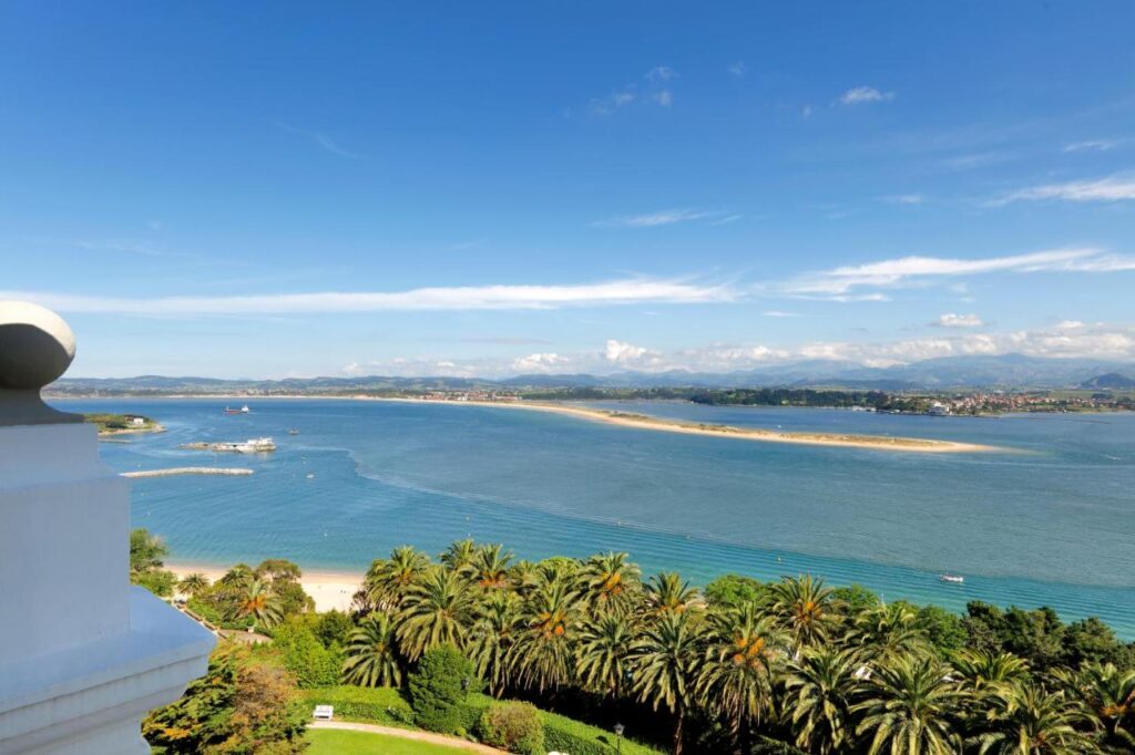 View from a high vantage point overlooking a tropical coastline with palm trees, sandy beach, blue ocean water, distant boats, and a town across the bay—reminiscent of scenic vistas near the best hotels in Cantabria under clear skies. Also one of the most unique hotels in Spain.