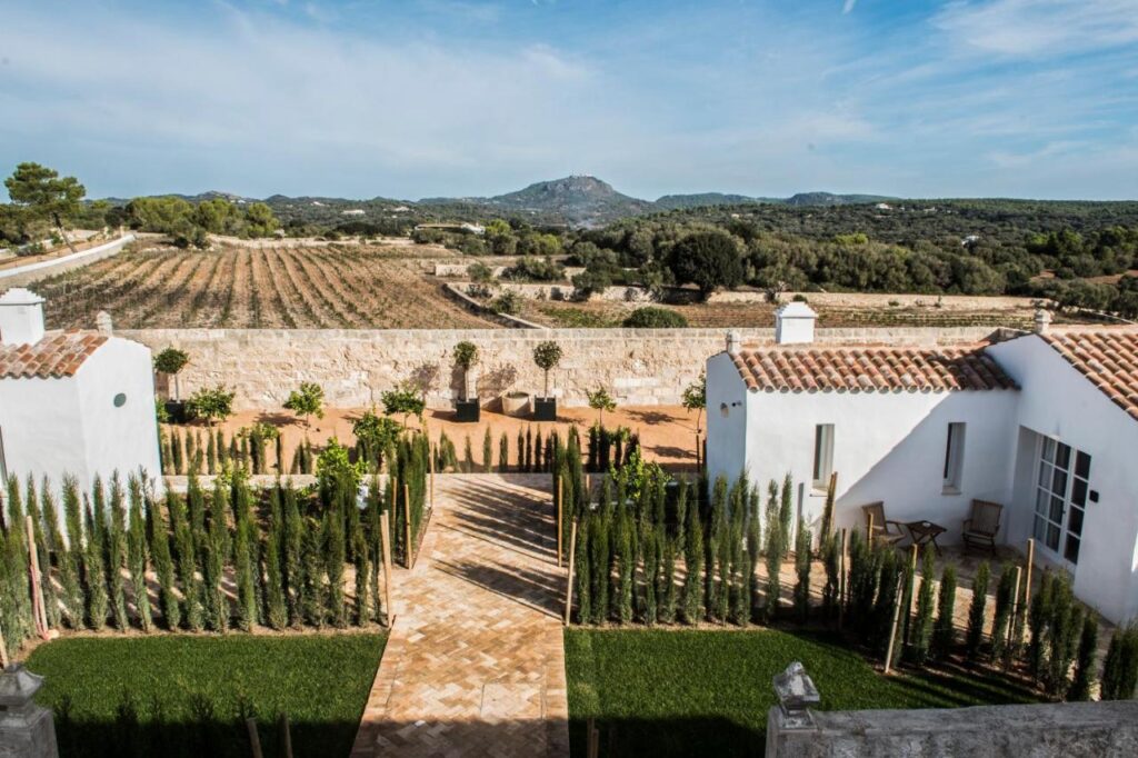 Unique Boutique Hotels in Menorca 26 Stone pathway lined with tall, narrow shrubs leads between two white, tile-roofed buildings—reminiscent of boutique hotels in Menorca—with fields, trees, and distant mountains under a partly cloudy blue sky.