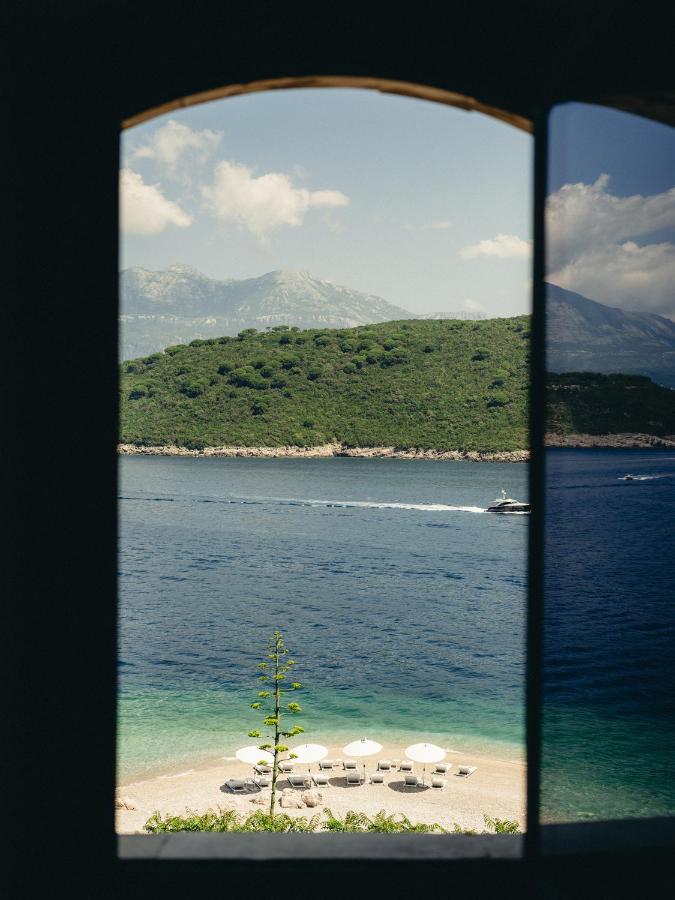 Most Authentic Boutique Hotels in Montenegro, Europe 3 View through an arched window at boutique hotels in Montenegro: a beach with white umbrellas, clear blue water, a green island, a passing boat, and mountains under a partly cloudy sky.
