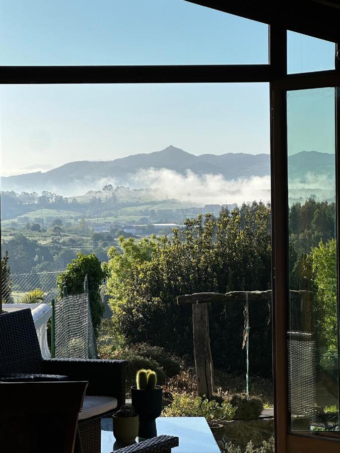 A scenic view from a window at hotels in Cantabria shows green hills, morning mist, and distant mountains under a clear sky. Patio furniture, plants, and a small cactus are visible in the foreground.