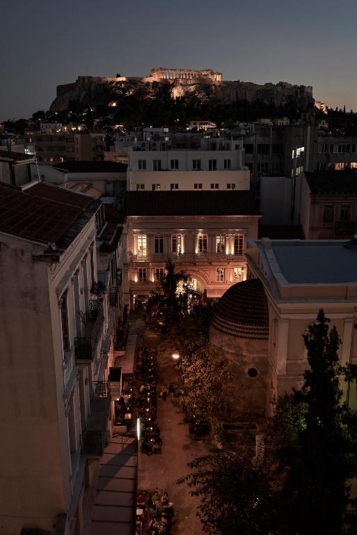 Nighttime view of the Acropolis illuminated on a hill, overlooking Athens city streets dotted with boutique hotels and outdoor dining areas, all aglow with warm lights below.