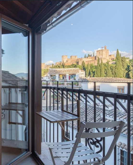 A small balcony at one of the boutique hotels in Granada features a white folding chair and a wooden table, overlooking tiled rooftops and the historic Alhambra palace surrounded by trees under a clear blue sky.