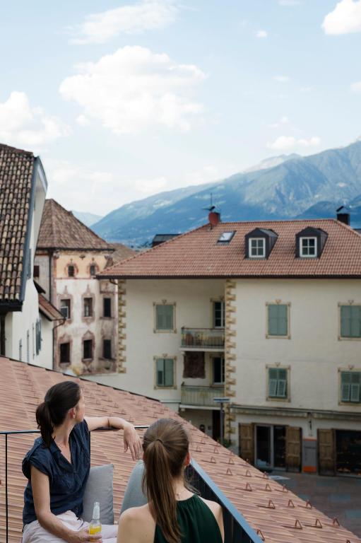 Two women sit on a rooftop terrace, looking out at traditional buildings and distant mountains under a partly cloudy sky. One woman holds a drink as they both face away, enjoying views reminiscent of the best hotels in the Dolomites.