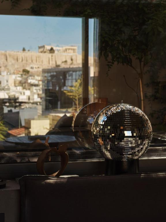 A shiny disco ball sits on a table indoors, with a large window reflecting light. Outside, the Acropolis and city buildings glimmer under a clear sky—offering views often found in boutique hotels in Athens.