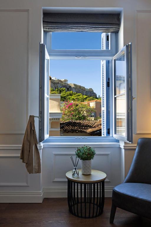 A bright room with an open window revealing a scenic view of the Acropolis, reminiscent of boutique hotels in Athens; blooming flowers and rooftops frame a small table with a plant and chair in the foreground.
