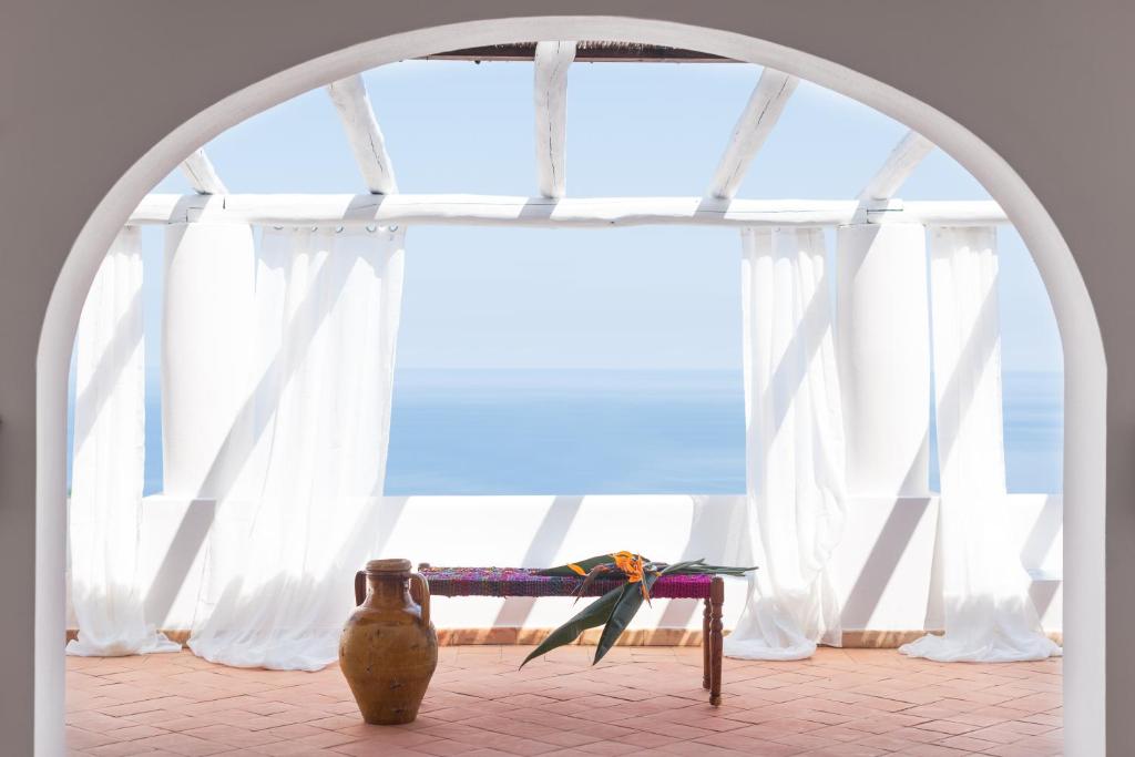 7 Best Hotels in the Aeolian Islands 7 A sunlit terrace in one of the best hotels in the Aeolian Islands, with white curtains, terracotta tiles, a rustic bench topped with a colorful cloth and tropical leaves, and a large clay jug overlooking a calm blue sea through an arched doorway.