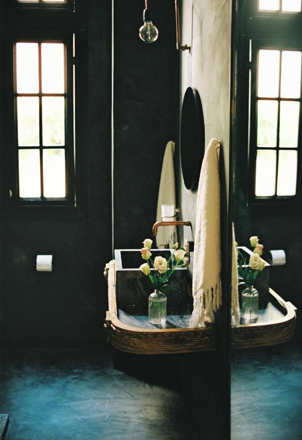 A bathroom with dark walls, two tall windows, a round mirror above a sink, and a vase of white roses evokes the elegance often found in the best boutique hotels in Athens. The scene is partially reflected in the mirror.