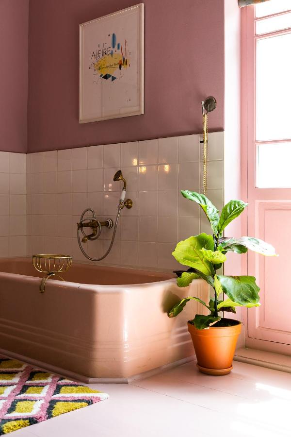 Unique Places to Stay in Cape Town 20 A pink-tiled bathroom with a matching pink bathtub, gold fixtures, a potted green plant, a colorful geometric rug, and framed artwork—a vibrant retreat among unique places to stay in Cape Town.