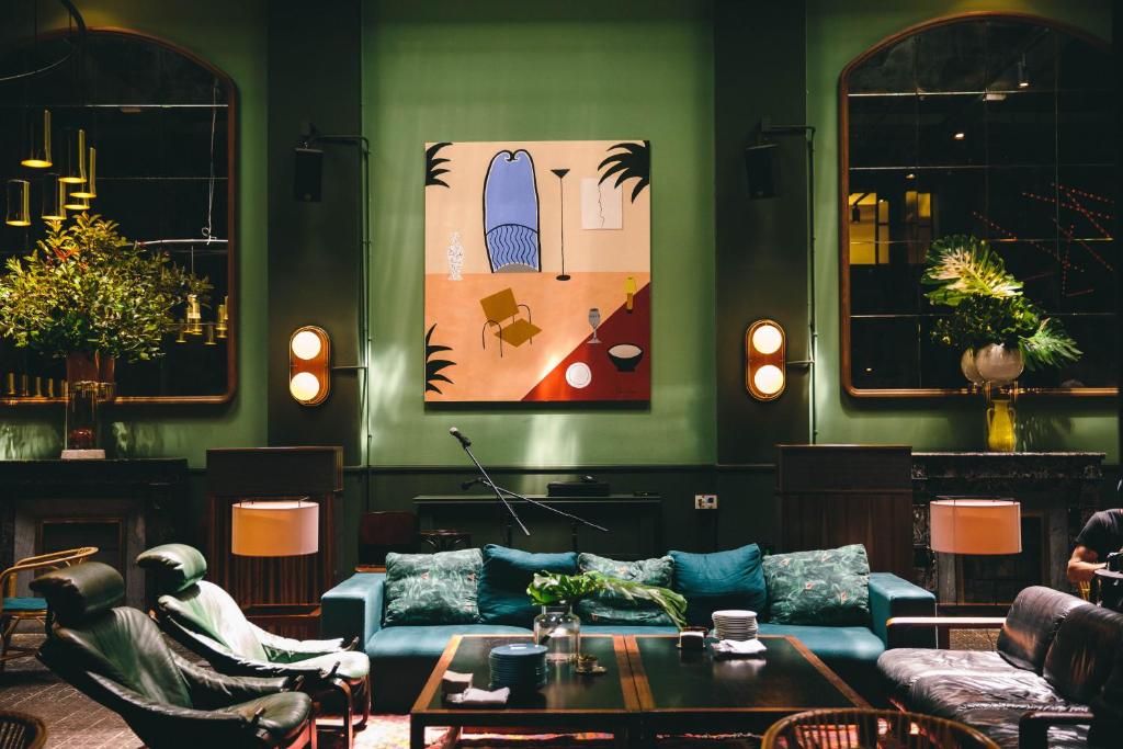 13 Unique Hotels in Barcelona, Spain 53 A stylish living room with green walls, a blue-green sofa, patterned cushions, and plants evokes the cozy, modern atmosphere often found in unique hotels in Barcelona. Large wall art, mirrors, and warm lighting complete the inviting space.