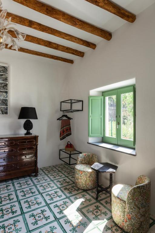 7 Best Hotels in the Aeolian Islands 13 A cozy room inspired by hotels in the Aeolian Islands, with wooden ceiling beams, patterned tile floor, floral armchairs, a dark wooden dresser with a black lamp, open shelves, and a window with green shutters welcoming natural light.