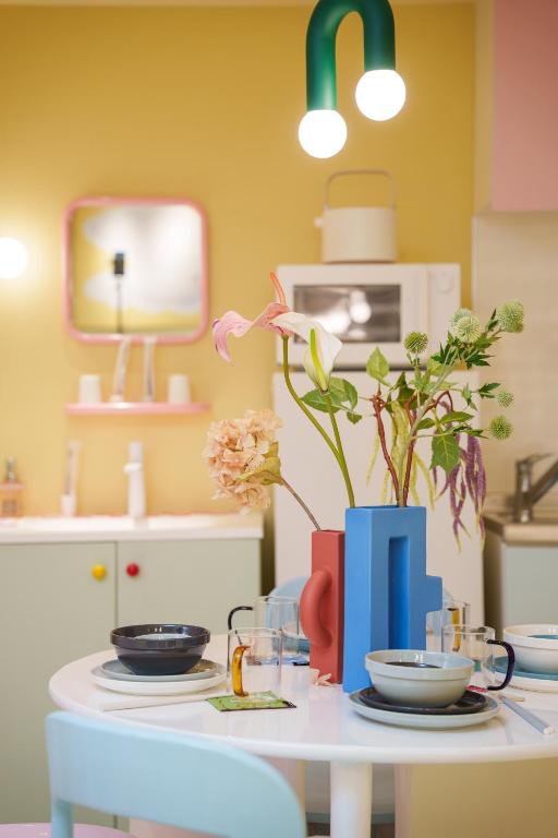 15 Unique Hotels in Tokyo: Coolest hotels 60 A modern, colorful kitchen with pastel cabinets, a white round table set with dishes, and a geometric blue and red vase holding flowers—reminiscent of the vibrant design found in unique hotels in Tokyo. Bright pendant lights create a cozy atmosphere.