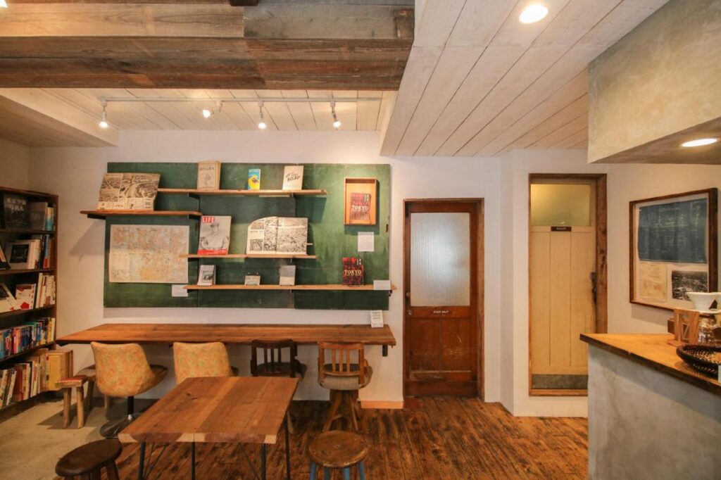 15 Unique Hotels in Tokyo: Coolest hotels 52 A cozy café with wooden tables and chairs, bookshelves, and art displayed on green wall shelves. Warm lighting creates an inviting atmosphere—perfect for those seeking the charm found in unique hotels in Tokyo.