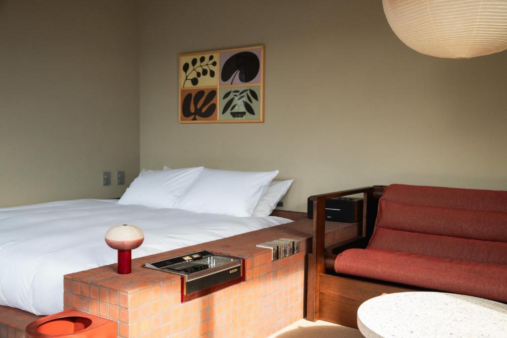 15 Unique Hotels in Tokyo: Coolest hotels 41 A modern bedroom inspired by unique hotels in Tokyo, featuring a white bed, a built-in brick nightstand with a round lamp, and a red cushioned sofa. Above the bed hangs framed abstract artwork as light streams in from the left.