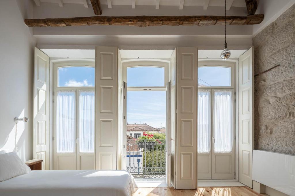 Top 12 Boutique Hotels in Porto, Portugal 41 A bright bedroom in the style of boutique hotels in Porto, with a bed on the left and three tall, white French doors opening to a balcony, letting in natural light and offering a view of rooftops and blue sky.