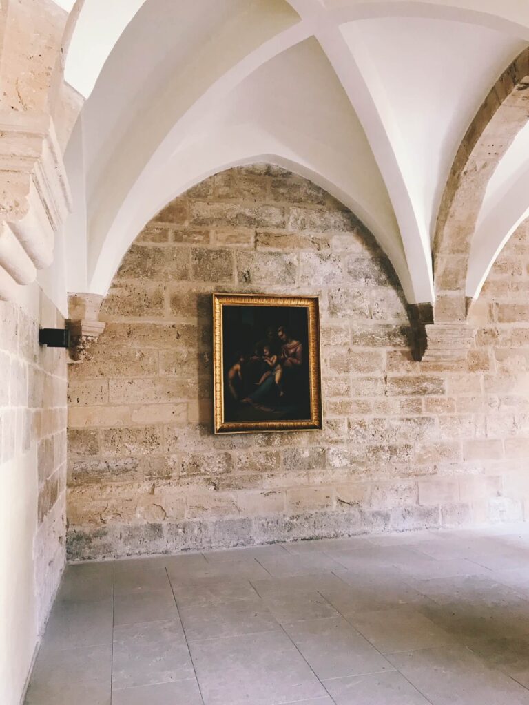 4 Best Hotels in Ribera del Duero, Spain 10 A framed painting hangs on a stone wall beneath high, vaulted ceilings with arched supports, evoking the elegant interiors found in exclusive hotels in Ribera del Duero. The spacious room features tile flooring and an inviting, tranquil ambiance.