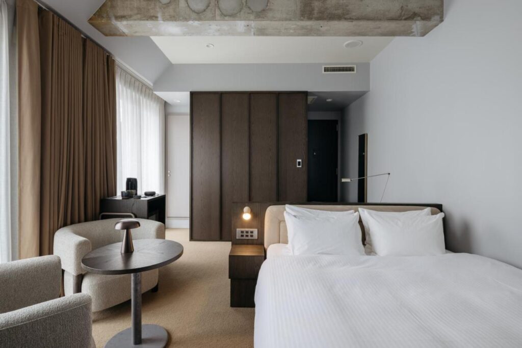 7 Best Hotels in Hiroshima, Japan 5 A modern hotel room, among the best hotels in Hiroshima Japan, featuring a large bed, two armchairs around a small round table, floor-to-ceiling windows with brown curtains, and minimalist decor in neutral tones.