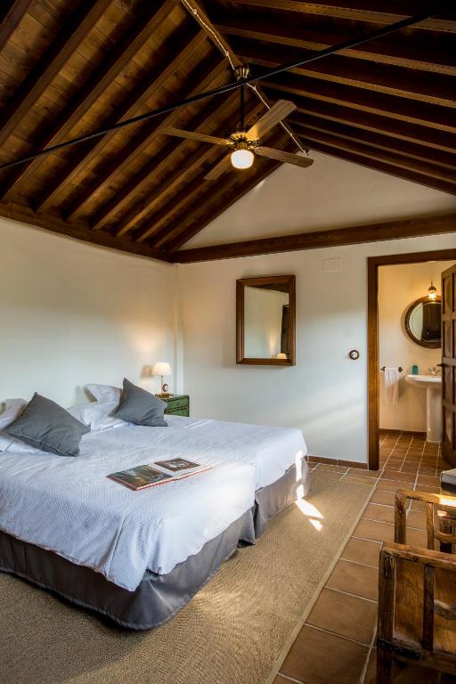 TOP 10 Luxury Hotels in Andalucía 31 A cozy bedroom inspired by luxury hotels in Andalucía, with twin beds, gray pillows, a wooden beamed ceiling, a mirror above a desk, a small lamp, and an open door to the bathroom. Warm natural light fills the rustic space.