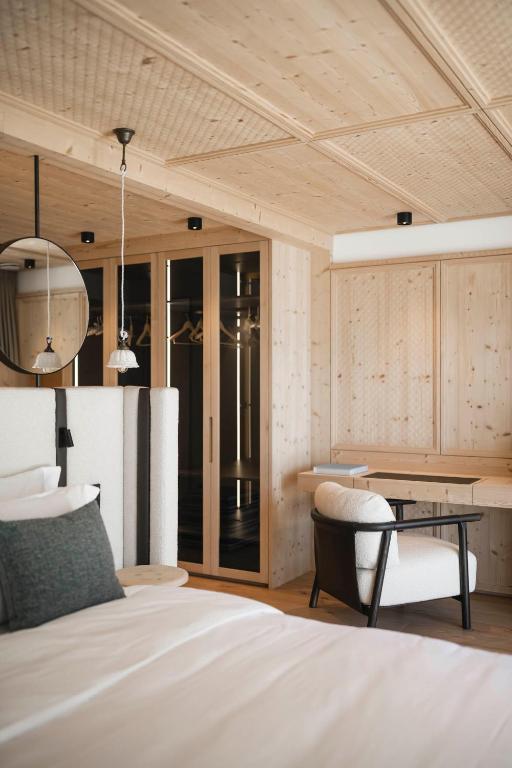A modern bedroom in the style of the best hotels in the Dolomites features light wooden walls and ceiling, a bed with white linens, a black and white armchair by a wooden desk, a round mirror, and an open closet with hanging clothes.