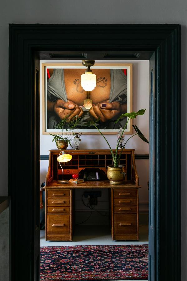 Unique Places to Stay in Cape Town 18 A wooden desk with plants and a lamp sits beneath a photo of a chest with tattooed lips, viewed through a dark doorway. Perfect for those seeking unique places to stay in Cape Town, with a patterned rug and ceiling light above.