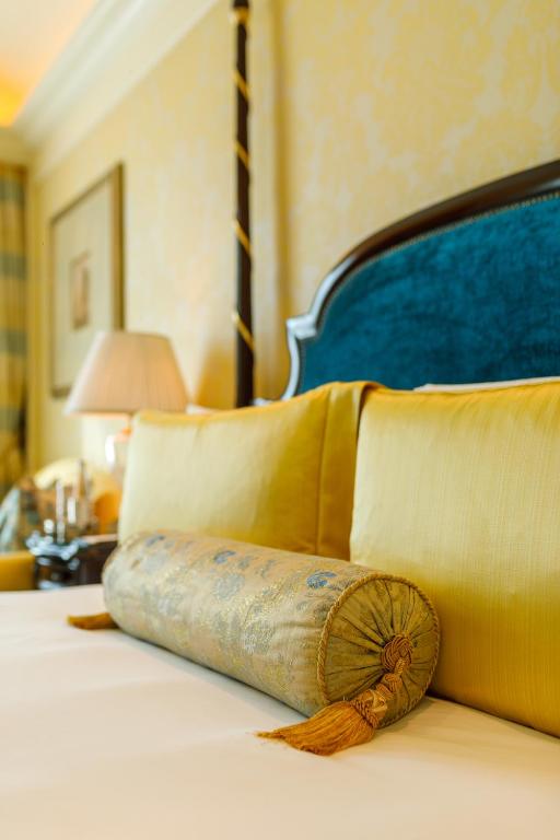 A close-up of a neatly made bed with yellow pillows and a decorative cylindrical cushion, set against a blue upholstered headboard—capturing the inviting ambiance found in boutique hotels in New Delhi. A lamp and framed picture adorn the softly lit bedside table.