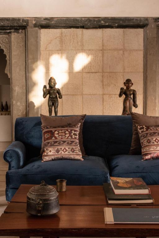 9 Iconic Heritage Hotels in Udaipur 38 A cozy living room, inspired by heritage hotels in Udaipur, features a blue velvet sofa, patterned pillows, bronze statues behind it, and a wooden coffee table with books and a decorative box. Warm sunlight casts soft shadows on the textured wall.
