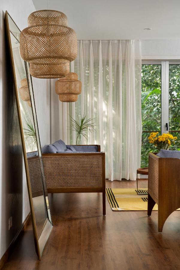 A modern living room reminiscent of boutique hotels in New Delhi, with wooden furniture, wicker pendant lights, a large floor mirror, sheer white curtains, a glass door overlooking the garden, and a wooden desk adorned with yellow flowers.