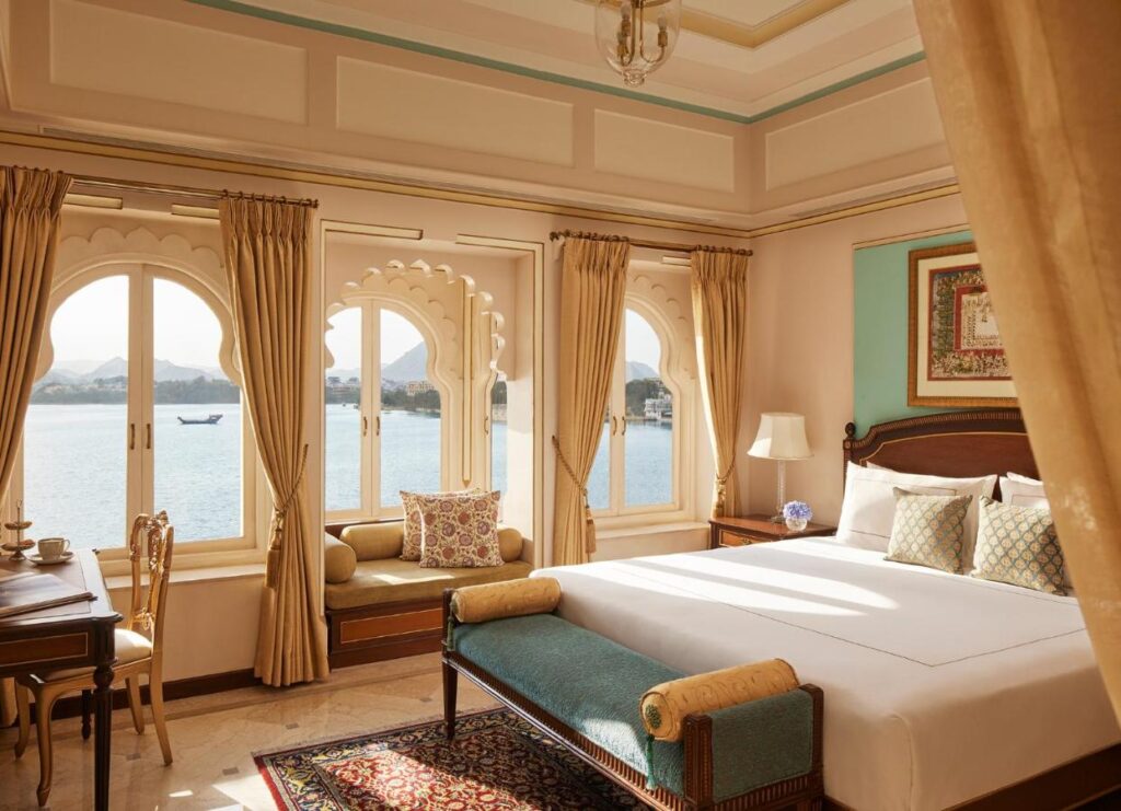 9 Iconic Heritage Hotels in Udaipur 12 Elegant hotel room in one of the heritage hotels in Udaipur, featuring a large bed, ornate windows overlooking a lake, golden curtains, cushioned window seat, desk, and classic warm-toned decor. Natural light fills the serene space.