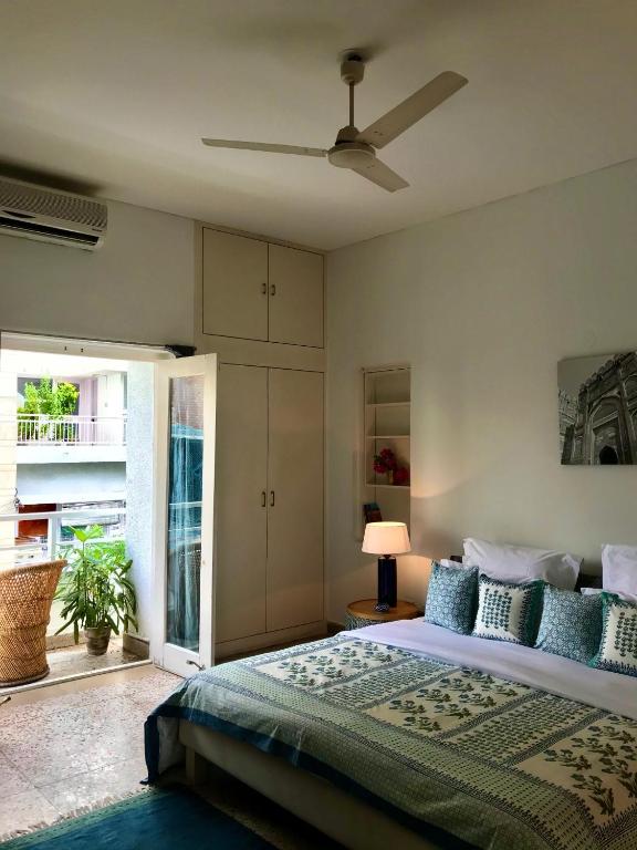 A bright bedroom inspired by boutique hotels in New Delhi, featuring a large bed with patterned bedding, a bedside lamp, wall art, ceiling fan, built-in wardrobe, air conditioning, and a door to a small outdoor balcony with plants.