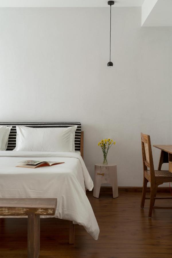 Minimalist bedroom inspired by boutique hotels in New Delhi: a bed with white linens, an open book, a side table with yellow flowers, a wooden chair, and a pendant light against a crisp white wall.