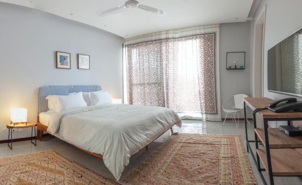A bright, modern bedroom inspired by boutique hotels in New Delhi, featuring a large bed with white bedding, side tables with lamps, patterned rugs, a wall-mounted TV, a chair, and large windows with patterned curtains letting in natural light.
