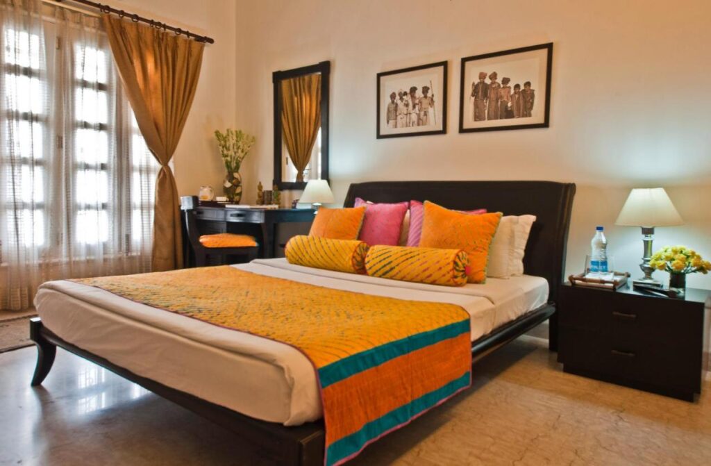 A cozy bedroom, inspired by boutique hotels in New Delhi, with a double bed featuring colorful pillows and a yellow-orange bedspread, bedside tables with lamps, a desk with a mirror, and framed art. Sunlight filters through beige curtains.
