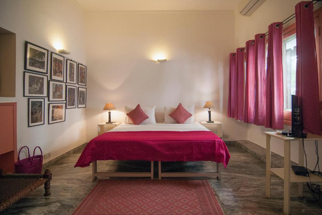 A cozy bedroom inspired by boutique hotels in New Delhi, featuring a double bed with a red bedspread and pillows, lamps on bedside tables, framed art, red curtains, a window, and a small desk with TV on a marble floor.